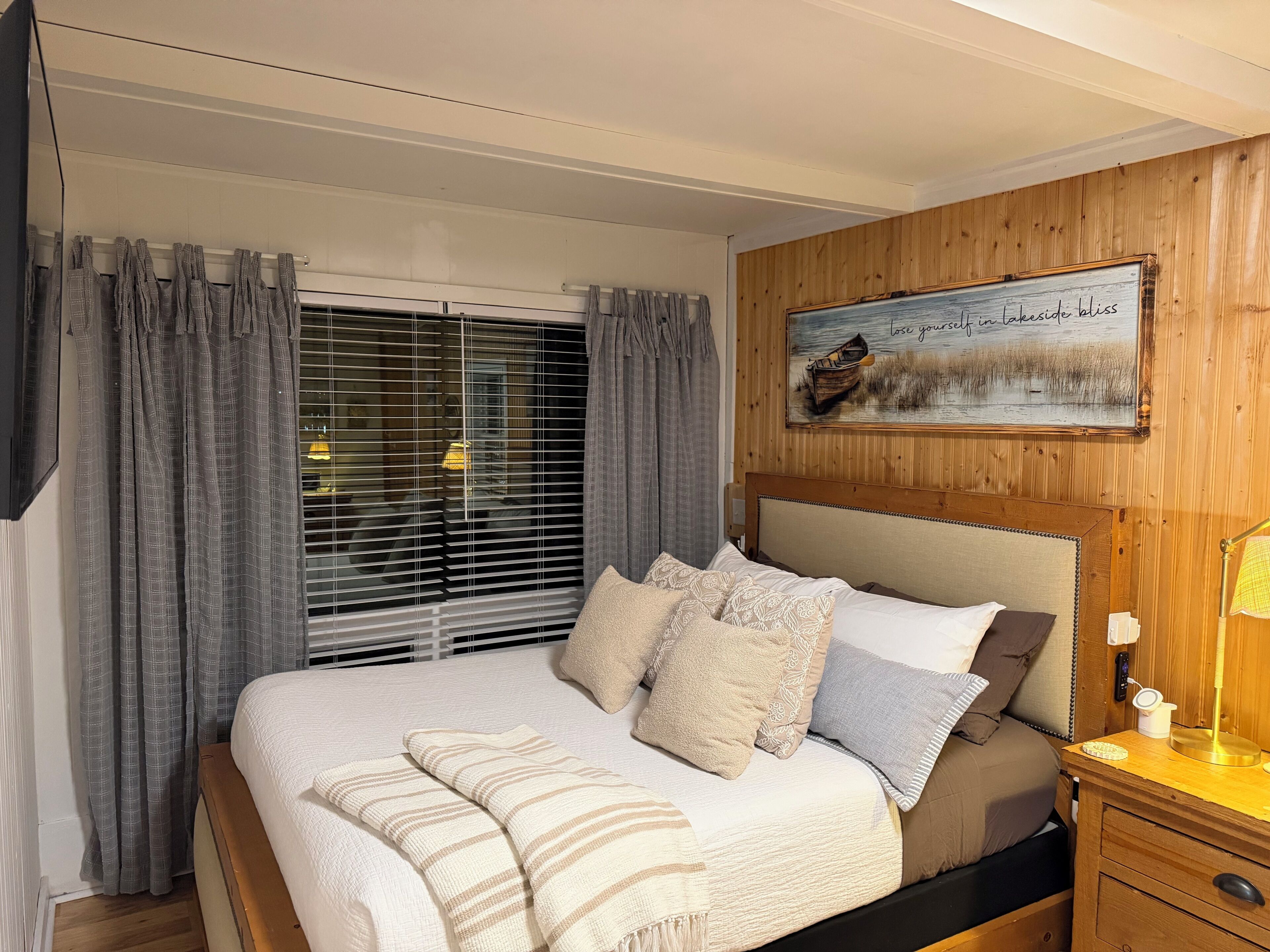 Une chambre à coucher comprenant un lit, des rideaux, une tête de lit, une table de chevet avec une lampe et un tableau accroché au mur.