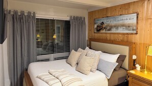 A bedroom with a bed, curtains, a headboard, a bedside table with a lamp, and a wall-mounted picture.