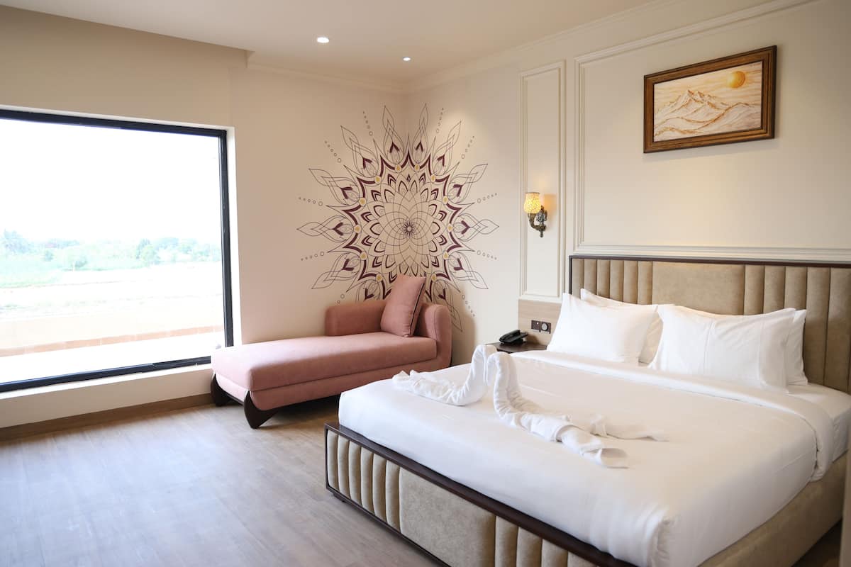 Habitación de hotel con un ventanal grande, cama con ropa blanca, un sillón rosa y un adorno en la pared.