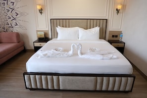 A neatly made bed with white linens and two towels arranged in a swan shape, a pink sofa, and a wall with a decorative pattern. - Vseven Hotels (Tiruvannamalai)