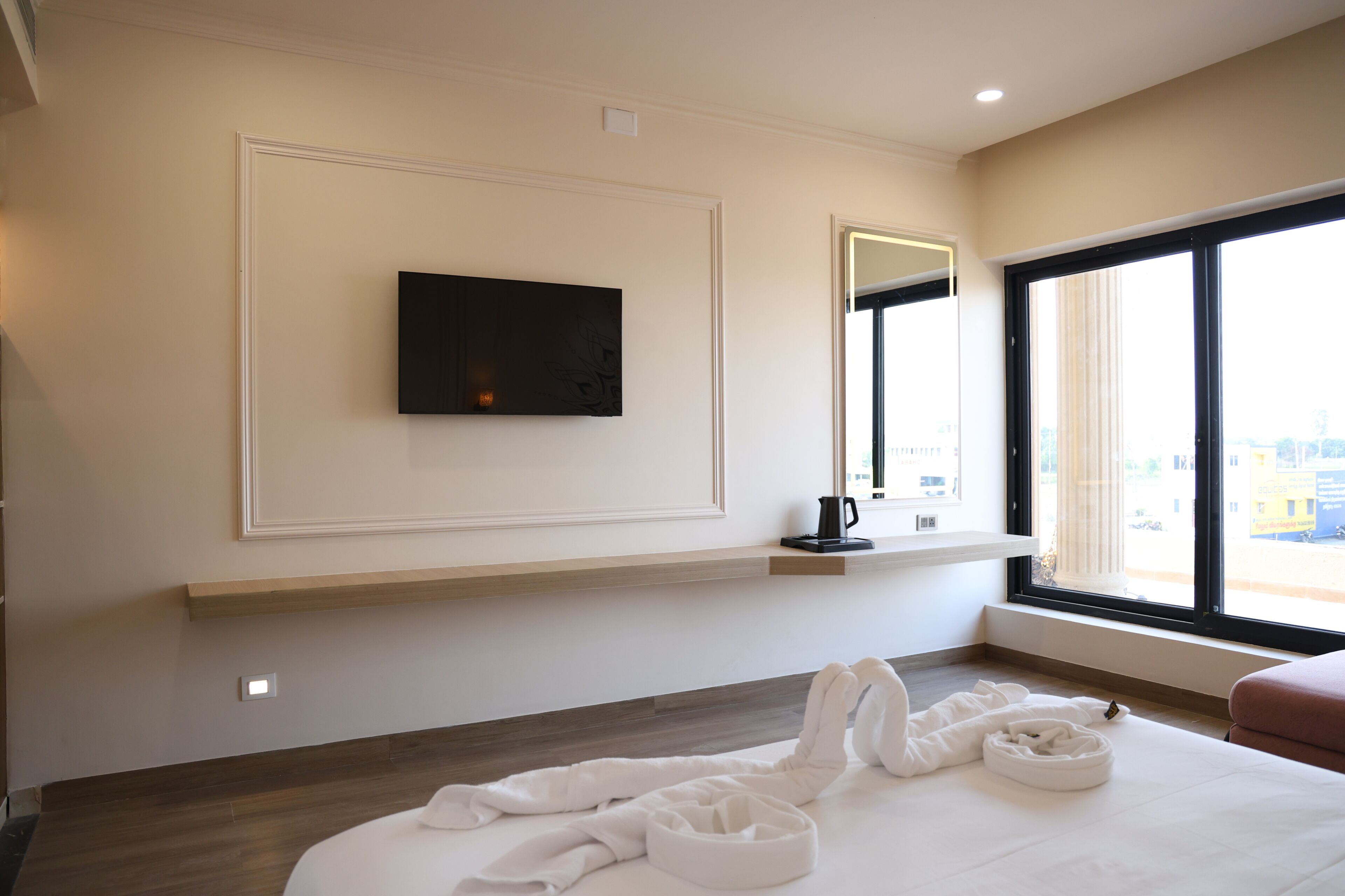 Habitación de hotel con televisión de pantalla plana montada en la pared, ventana con vistas al exterior, piso de madera y cama con ropa blanca.