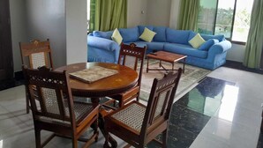 Een woonkamer met een blauwe bank, groene gordijnen, een ronde houten tafel met stoelen en een betegelde vloer.