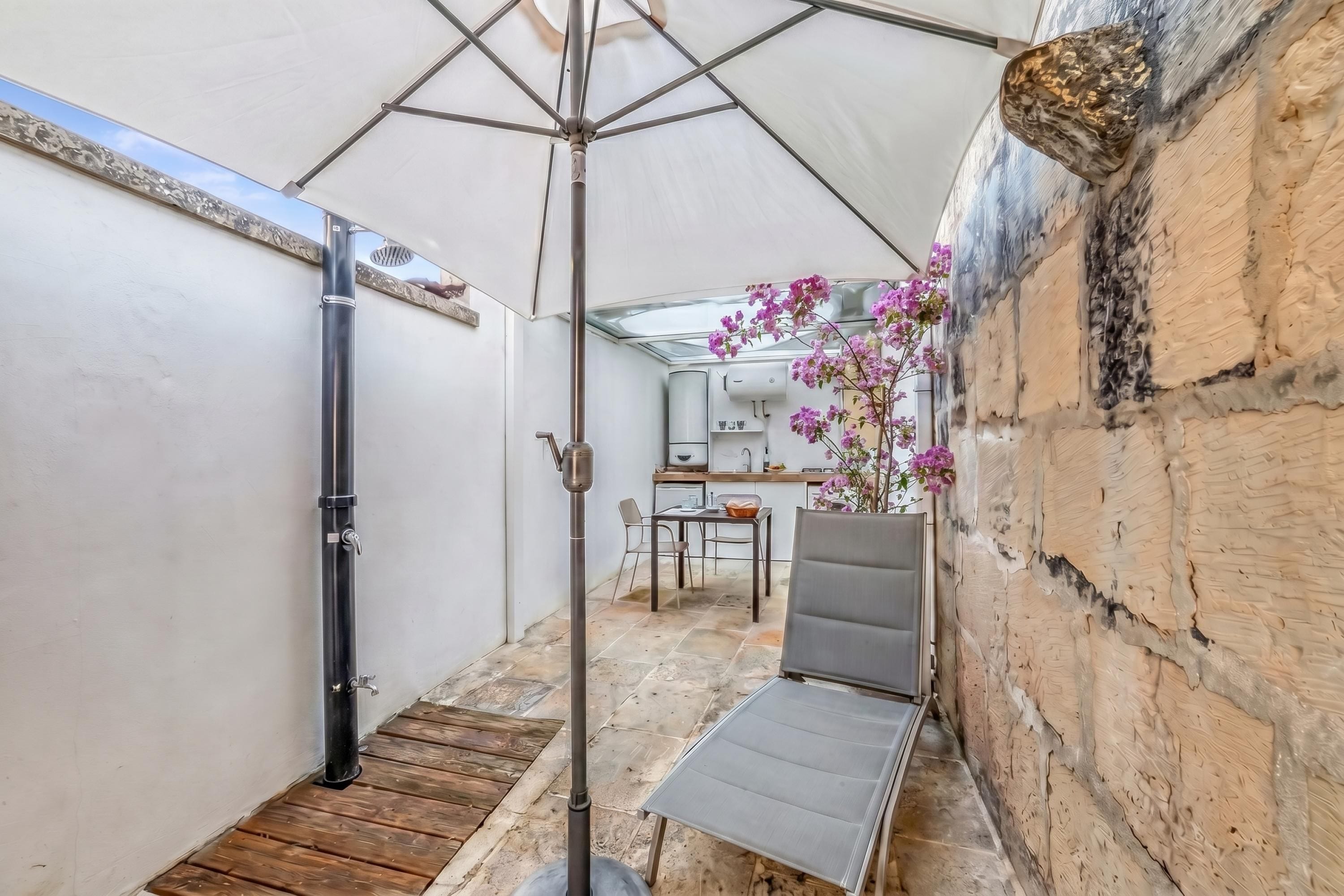 En smal udendørs terrasse med en metal parasol, en træbænk, et bord og et køkkenområde i baggrunden.