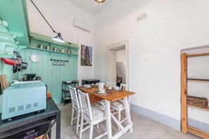 Un coin repas avec une table en bois et des chaises blanches, un espace cuisine avec un appareil électroménager bleu, et une étagère fixée au mur où sont rangés les ustensiles de cuisine.