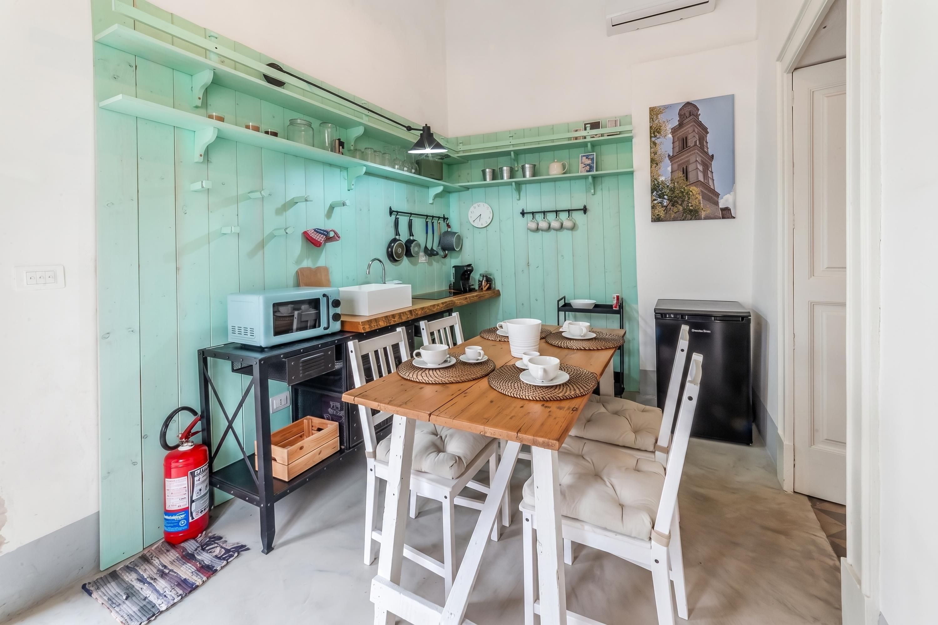 Cocina compacta con paredes color turquesa, una mesa de comedor de madera, sillas blancas y un refrigerador negro.