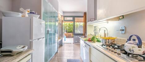 A modern kitchen with a refrigerator, oven, and stove. There is a window providing natural light and a view of the outdoors.