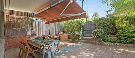 An outdoor patio with a table and chairs, a covered area, and a garden with plants and a fence.