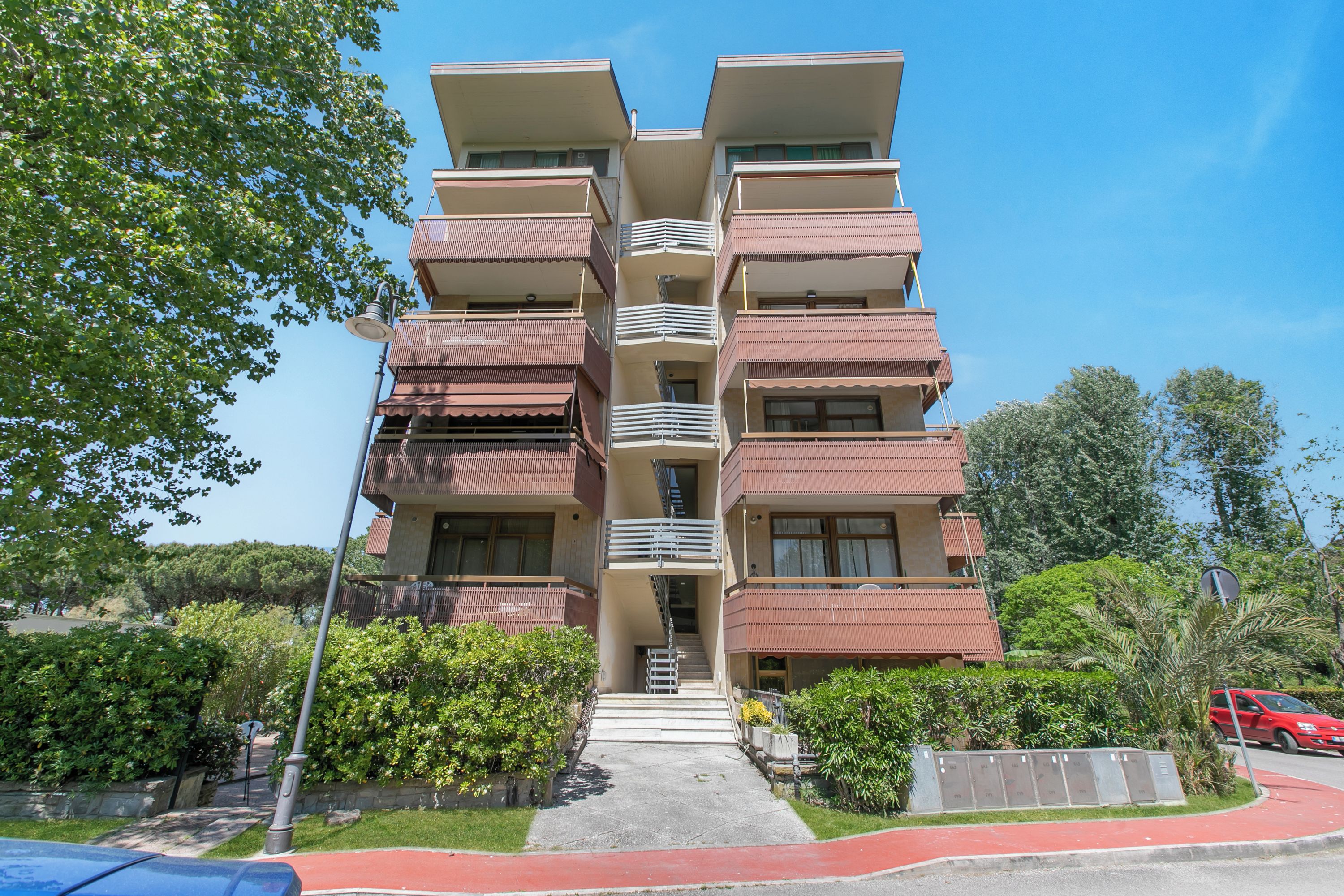 A multi-story residential building with balconies, a central staircase, and a red pathway leading to the entrance.