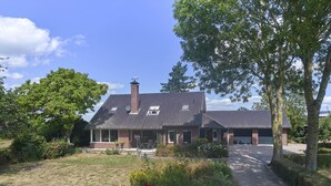 Une maison à deux étages, avec une façade en briques, un toit en tuiles et un porche à l’avant, agrémenté d’un banc.