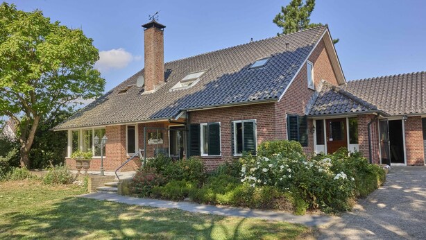 Une maison à deux étages, construite en briques, avec un toit en pente, une cheminée et un porche à l’avant, doté d’une balustrade.