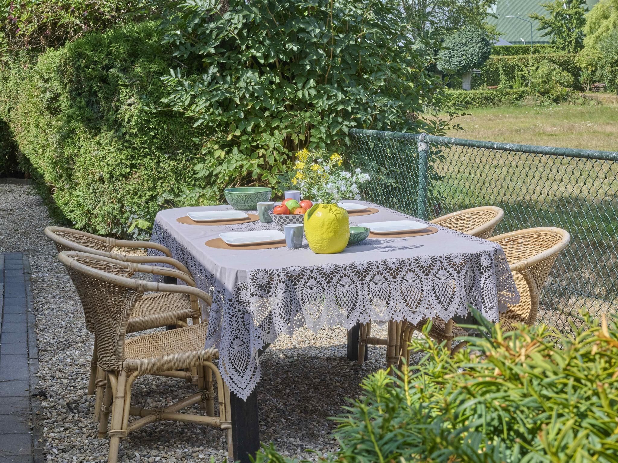 Et udendørs spiseområde med et bord dækket til fire personer, omgivet af fletstole, en hvid dug og en gul vase med blomster.