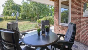 Une terrasse extérieure avec une table ronde, deux chaises, un pot de fleurs et un panneau.