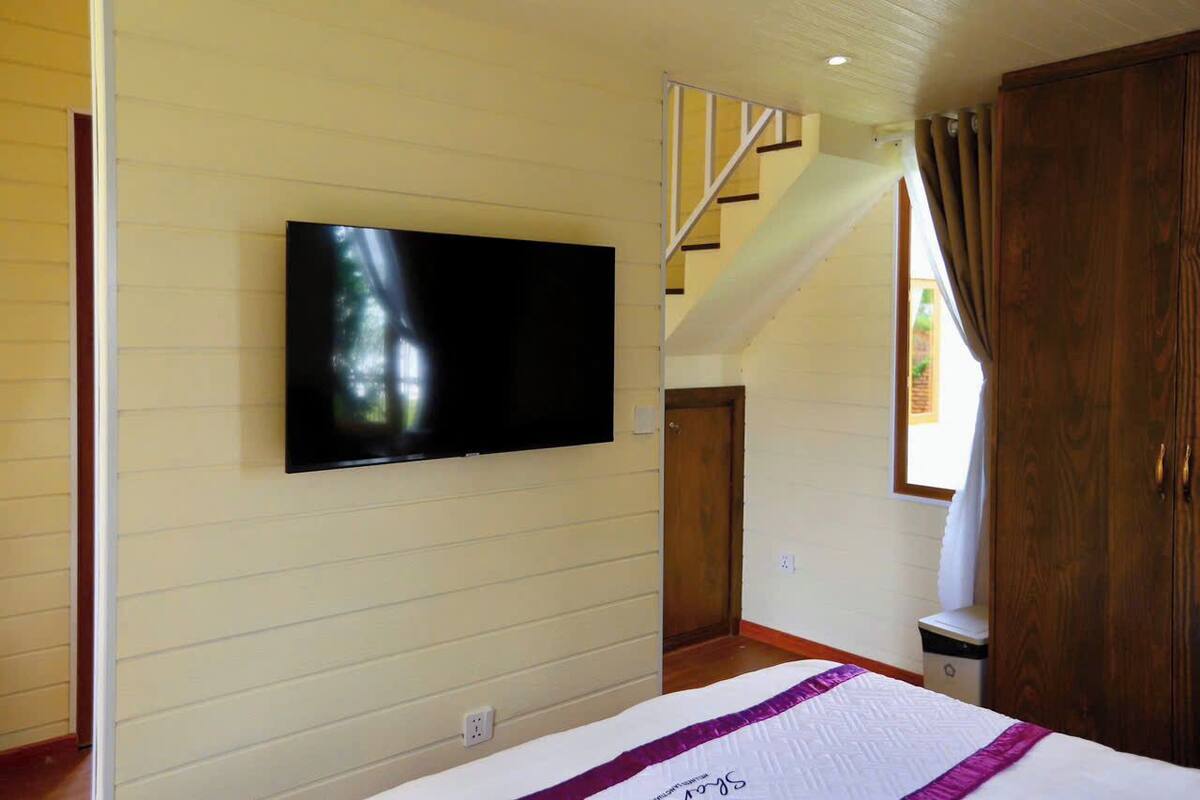 A bedroom with a bed, a television mounted on the wall, a staircase, and a window with curtains.