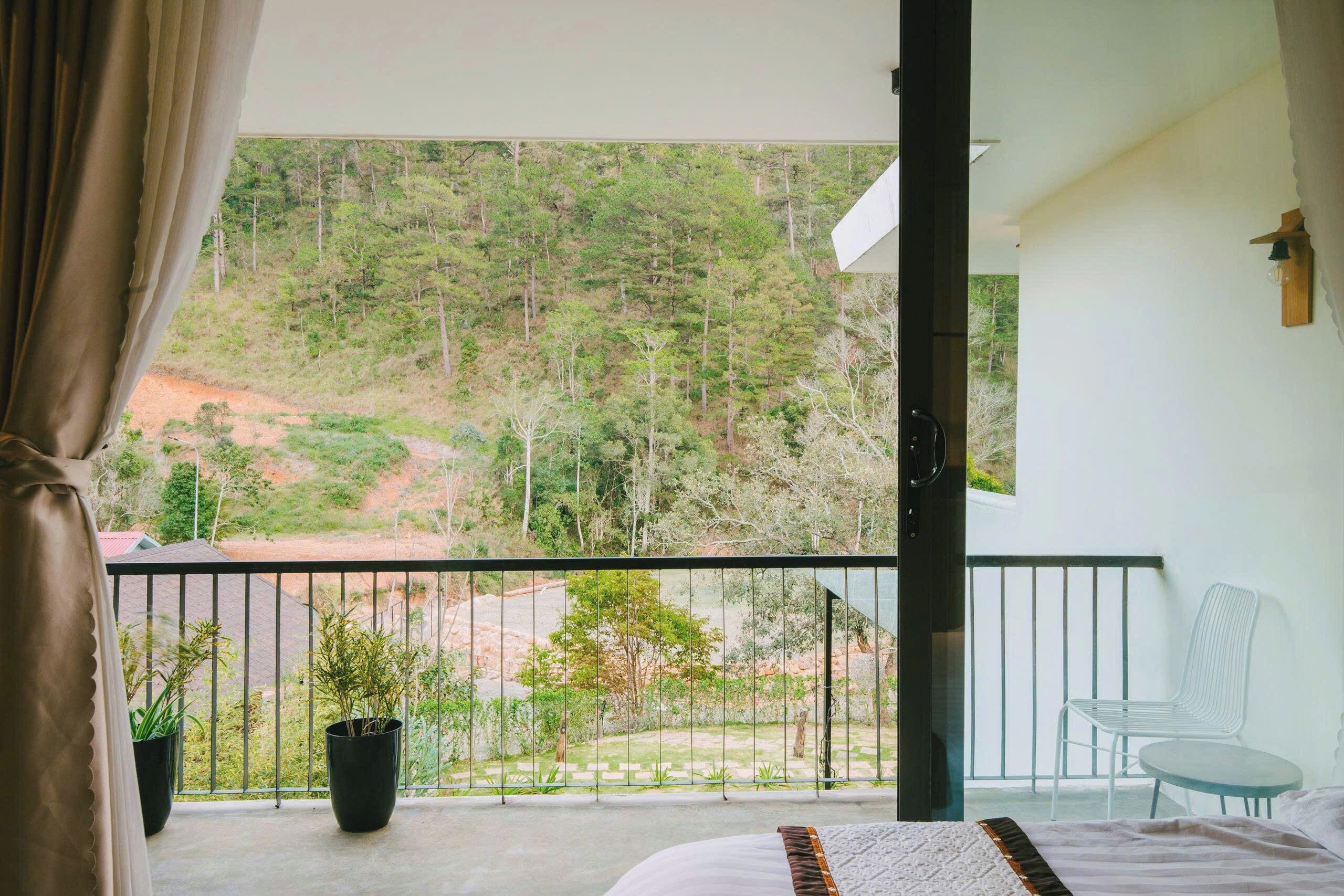 Un balcón con vistas a una ladera boscosa, una cama con edredón blanco y marrón, una silla blanca y una planta en maceta.