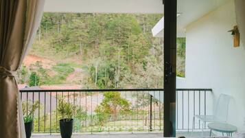 Un balcón con vistas a una ladera boscosa, una cama con edredón blanco y marrón, una silla blanca y una planta en maceta.