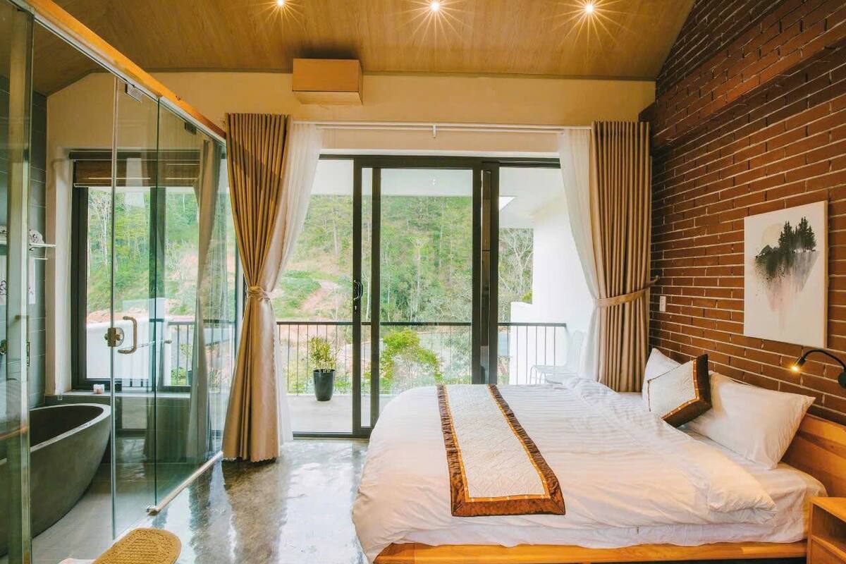A bedroom with a large bed, a glass shower enclosure, and a balcony with a view.