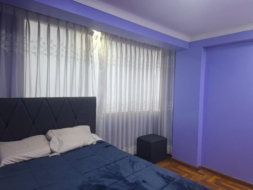 A bedroom with a bed, a small stool, and a window with sheer curtains.