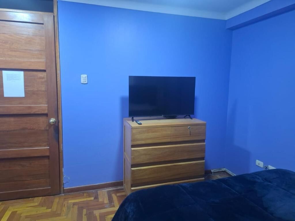 A room with a wooden door, a blue wall, a wooden dresser with a flat-screen TV on top, and a bed with a dark cover.