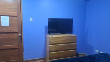 A room with a wooden door, a blue wall, a wooden dresser with a flat-screen TV on top, and a bed with a dark cover.