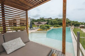 Área à beira da piscina com pérgola de madeira, espaço de estar com almofadas e vista para a piscina e vegetação ao redor.
