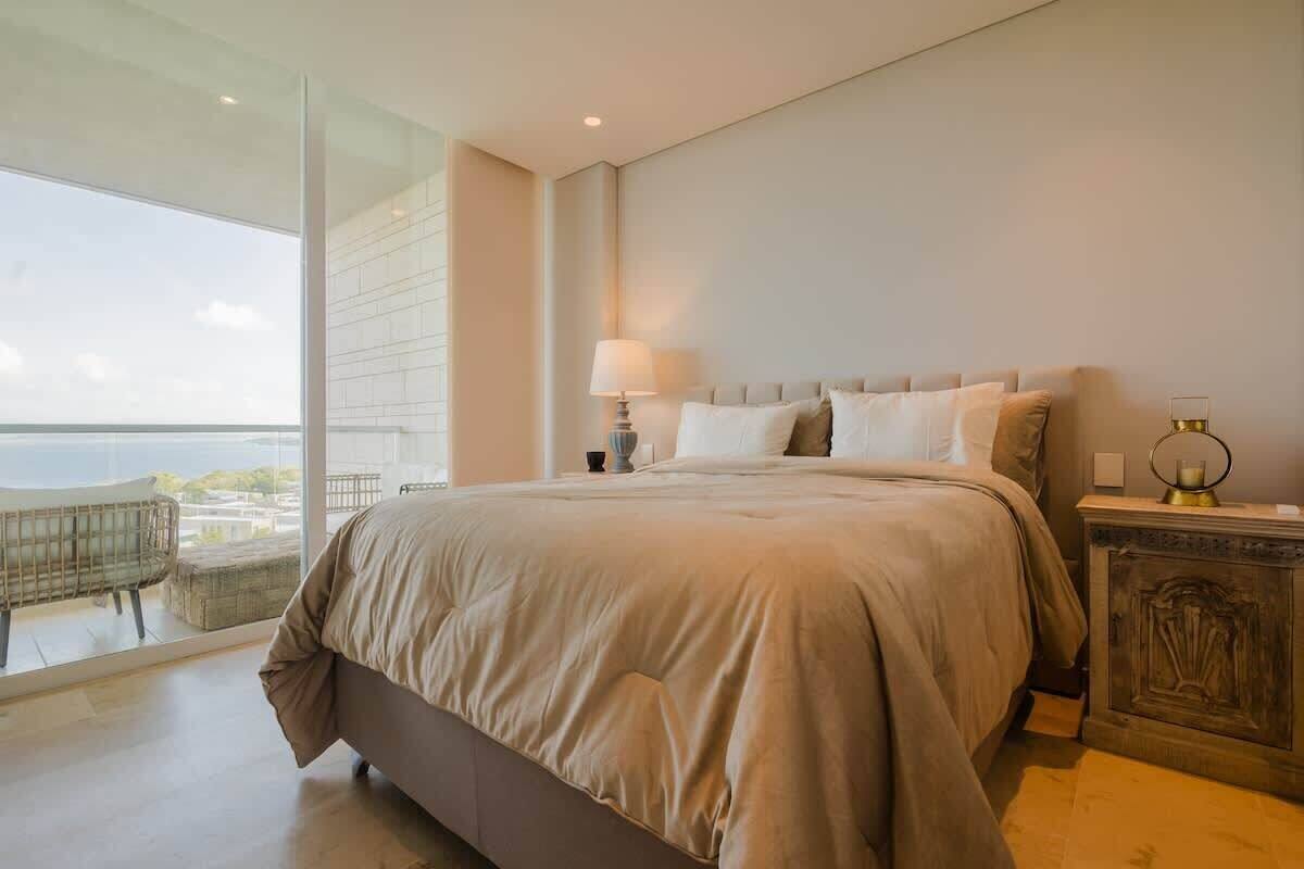 A bedroom with a large bed, a nightstand, a lamp, and a view of the outdoors.