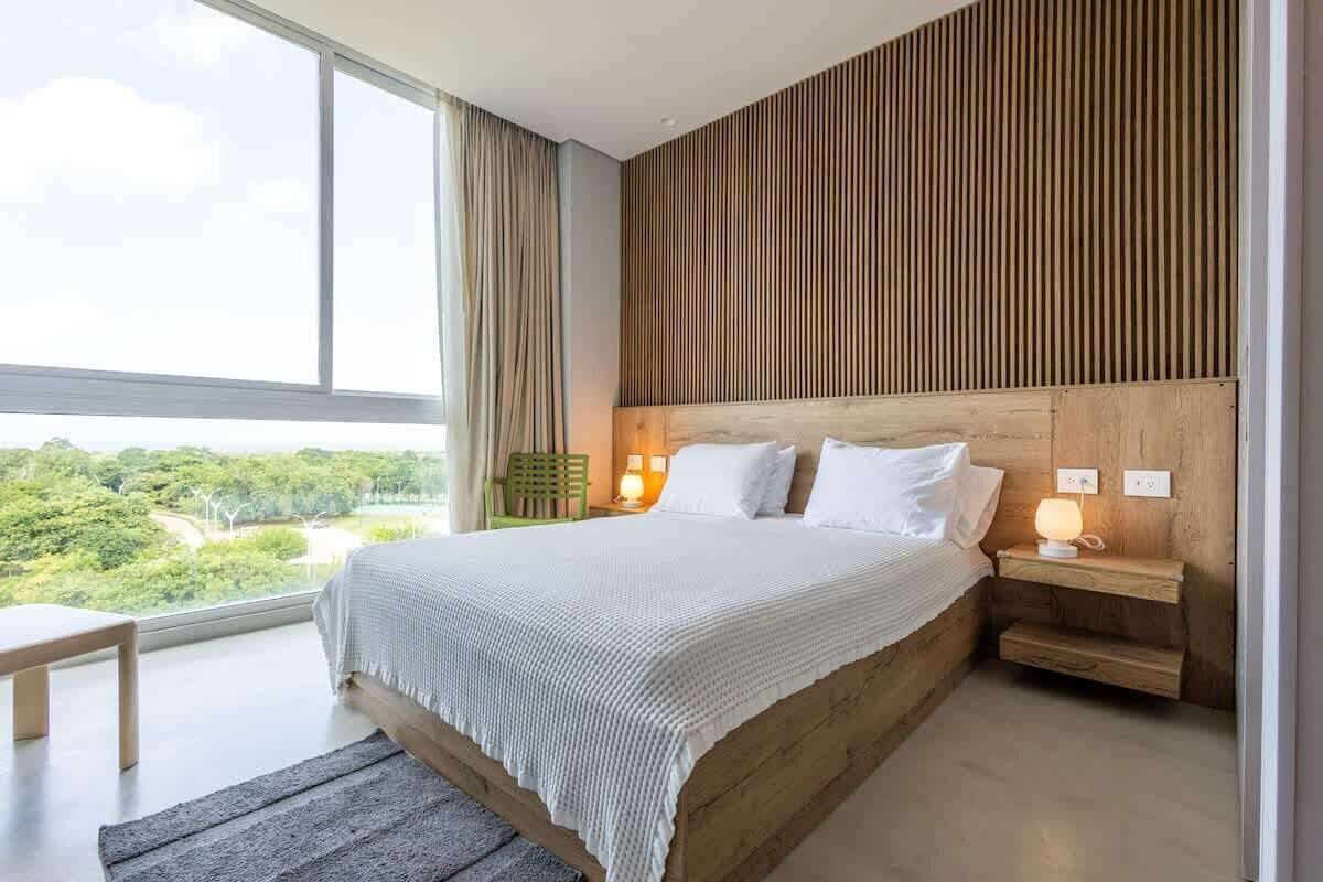 A modern bedroom with a large bed, wooden headboard, bedside tables, and a view of greenery outside.
