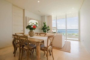 Un comedor con mesa y sillas de madera, un florero con flores y vistas al mar a través de amplios ventanales.