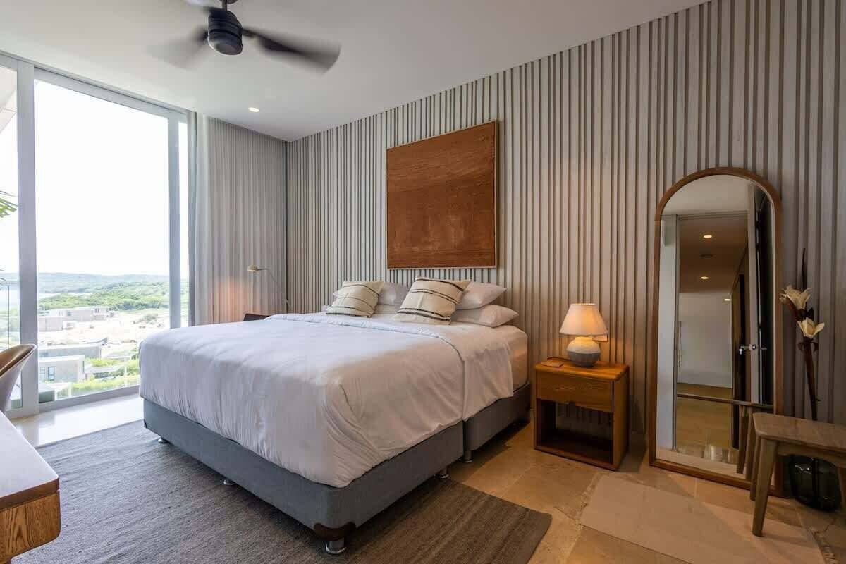 A modern bedroom with a large bed, a bedside table with a lamp, a mirror, and a view of the outdoors.
