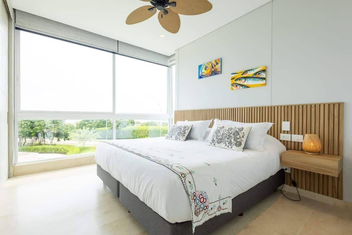 A modern bedroom with a large bed, a wooden headboard, a nightstand with a lamp, and artwork on the wall.