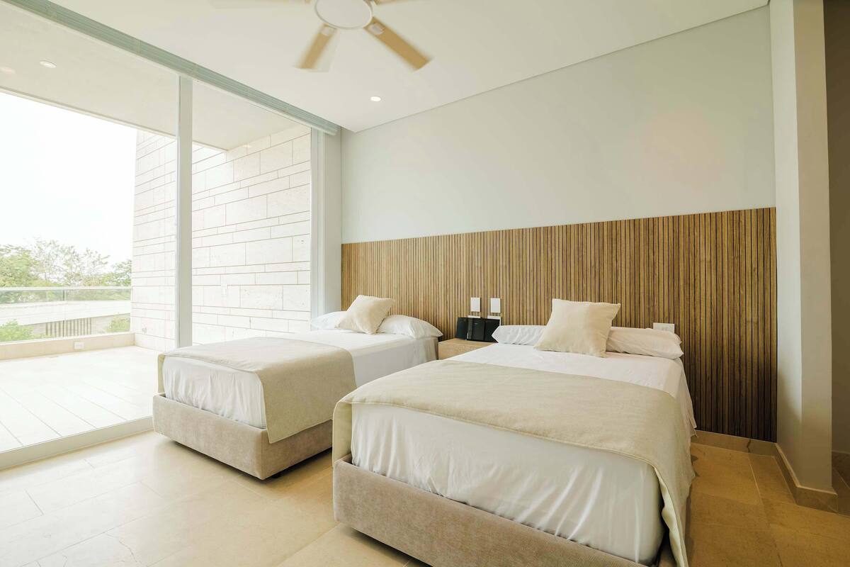 A modern hotel room with two beds, a large window, and a ceiling fan.