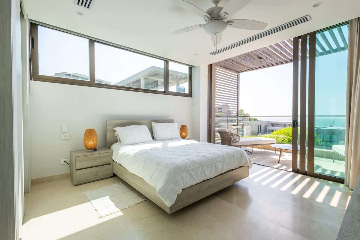 A modern bedroom with a large bed, a nightstand, a ceiling fan, and a view of the outdoors through large windows.