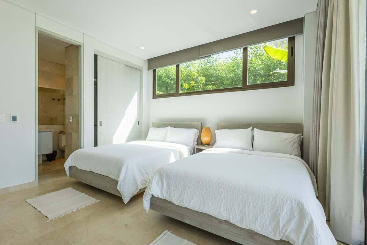 A modern bedroom with two beds, a large window, and a view of greenery.
