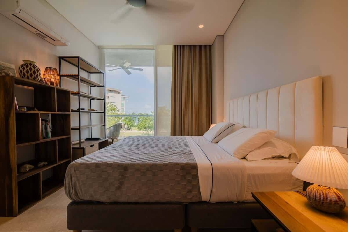 A modern bedroom with a large bed, a wooden nightstand, a lamp, and a bookshelf.