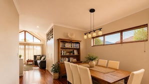 A dining area with a wooden table, white chairs, a bookshelf, and a window with blinds. - Bay Breeze on Thomas (West Busselton)