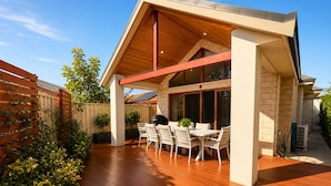 A covered patio with a table and chairs, a wooden deck, and a brick wall. - Bay Breeze on Thomas (West Busselton)