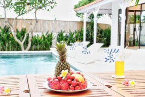 A poolside setting with a fruit bowl, a pineapple, and a drink on a wooden table. - Specious 3Bedroom Rakuen Villa Canggu (Canggu)