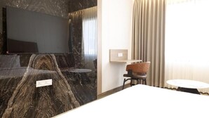 A modern hotel room with a large flat-screen TV mounted on a marble wall, a wooden chair, a small round table, and a bed with a white headboard.
