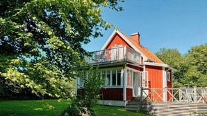 Ett rött trähus med en balkong, omgivet av en gräsklädd trädgård och träd.