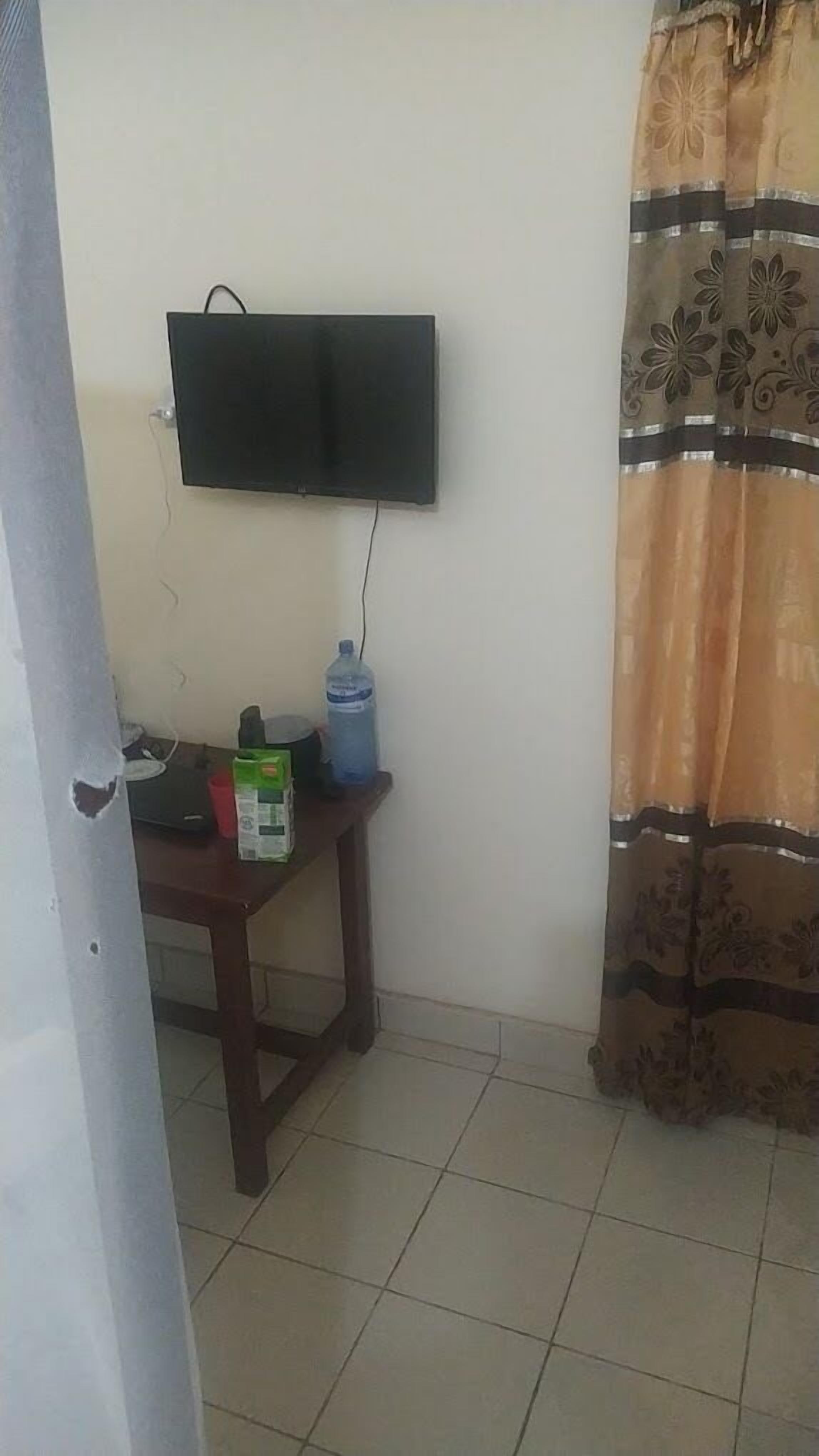 A room with a flat-screen TV mounted on the wall, a water dispenser, a small table with a green box, and a patterned curtain.