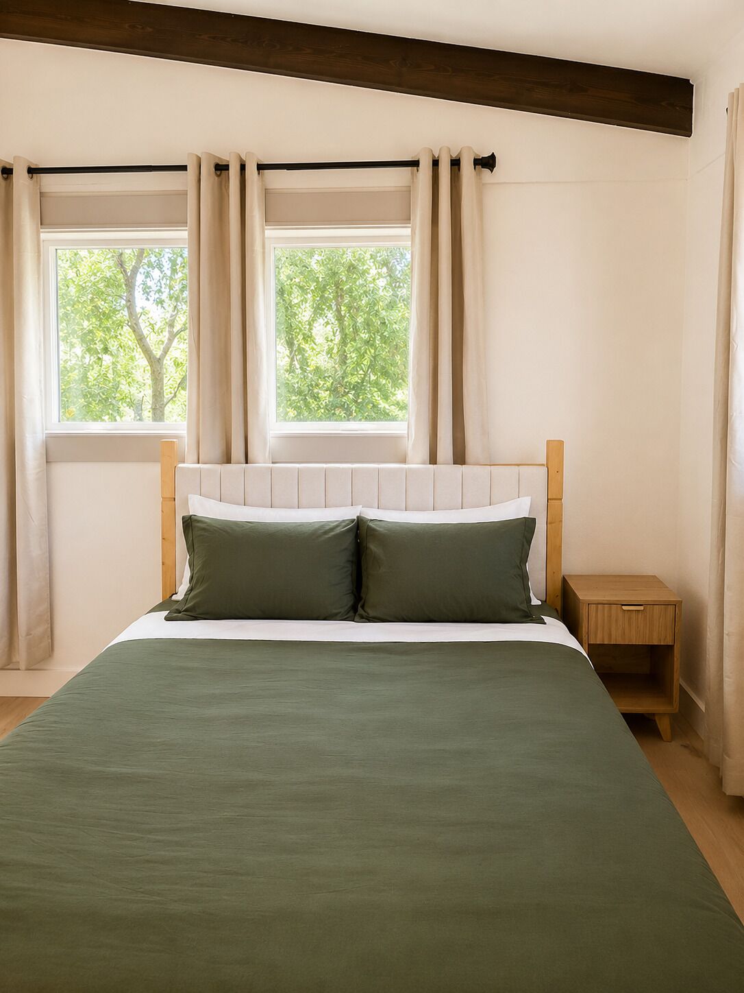 A bedroom with a bed, two windows with curtains, a wooden headboard, and a nightstand.