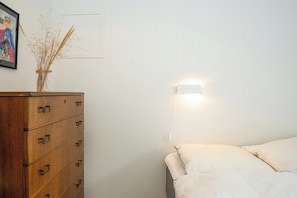 A bedroom with a wooden dresser, a bed with white bedding, a wall-mounted light, and a framed picture above the dresser. - Summer house in Houstrup with scenic views (Nørre Nebel)