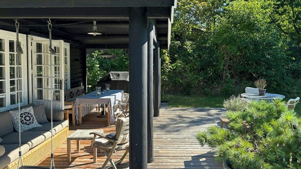 A covered outdoor dining area with a wooden deck, wicker furniture, and a swing chair. - Log cabin on beautiful natural grounds in Hornbæk (Hornbæk)