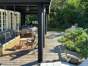 A covered outdoor dining area with a wooden deck, wicker furniture, and a swing chair. - Log cabin on beautiful natural grounds in Hornbæk (Hornbæk)