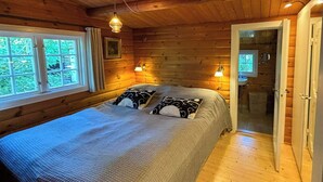 A wooden cabin bedroom with a bed, two bedside lamps, a window, and a door leading to a bathroom. - Log cabin on beautiful natural grounds in Hornbæk (Hornbæk)