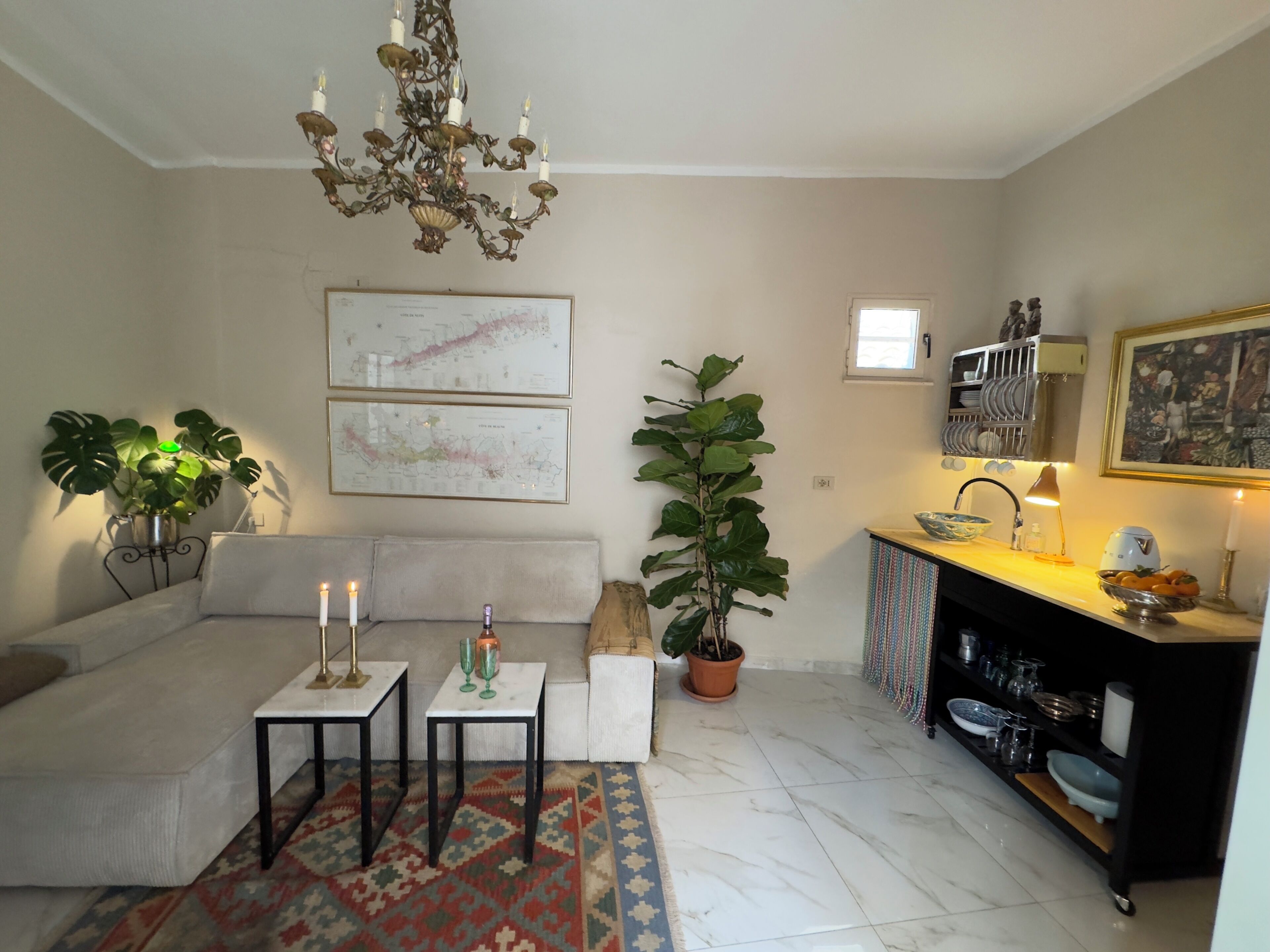 Una sala de estar con sofá, una mesa de centro, una araña, una planta en maceta, un comedor con un fregadero, y dos mapas enmarcados en la pared.