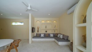 Ein modernes Wohnzimmer mit einem Ecksofa, einem Esstisch mit Stühlen, einem Deckenventilator, Einbauleuchten und Regalen mit dekorativen Gegenständen.
