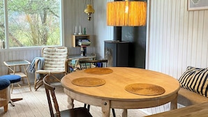 A wooden dining table with woven placemats, a wicker chair, a cushioned bench, and a black stove in the background. - Summer house near beach in Odder (Odder)