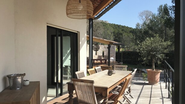 Eine Terrasse im Freien mit einem Holztisch und -stühlen, einer hängenden Laterne und Blick auf einen bewaldeten Hügel.