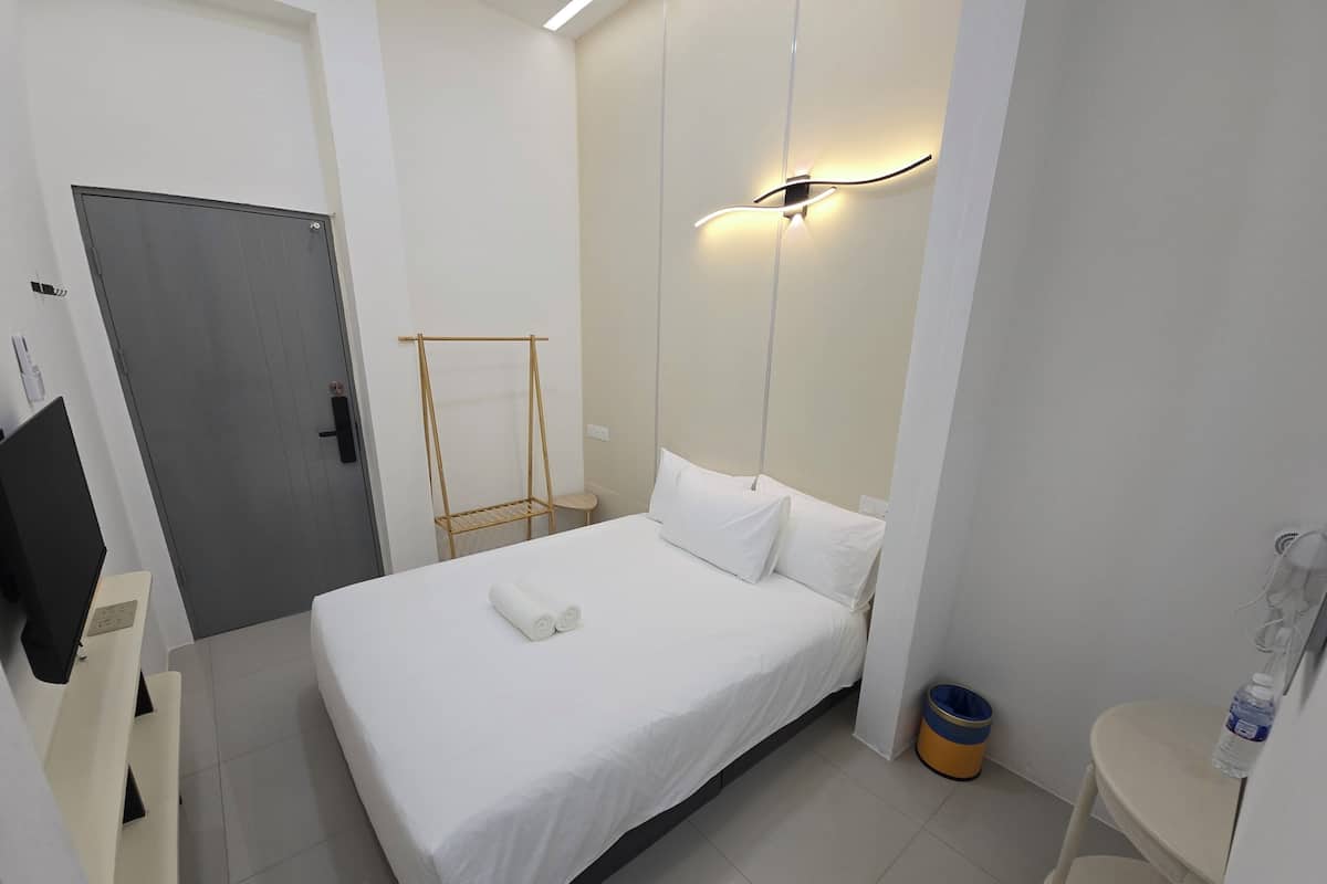 Quarto de hotel com cama, mesa de cabeceira, um banquinho, arandela, porta e lixeira.