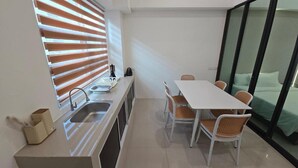 Cocina compacta con lavamanos, una pequeña encimera y un ventanal con persianas. Se observa una mesa de comedor con cuatro sillas y una cama con colcha verde visible a través de una puerta de cristal.
